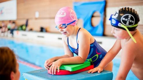 Junge Schwimmerin mit rosafarbenen Badekappe und Schwimmbrett am Beckenrand, beobachtet von einem Jungen.