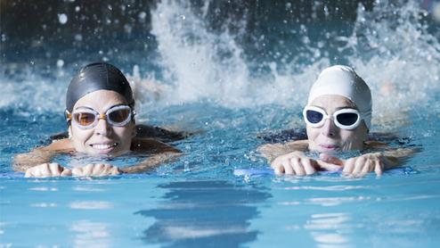 Zwei Schwimmerinnen in Badekappen üben mit Schwimmhilfen in einem beheizten Becken, Spritzer umgeben sie.