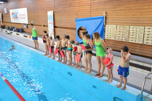 Gruppe von Schwimmschülern und Lehrern am Beckenrand eines Hallenbads, einige halten Schwimmhilfen.
