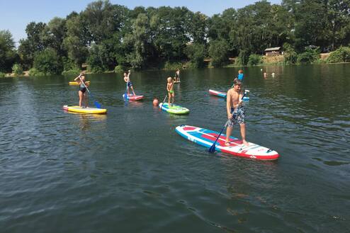 Fünf Kinder paddeln auf Stand-Up-Paddle-Boards auf einem ruhigen Gewässer unter blauem Himmel.