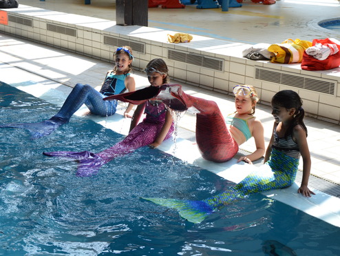 Vier Kinder mit flüssigen Schwimmkostümen sitzen am Beckenrand eines Schwimmbads und spielen im Wasser.