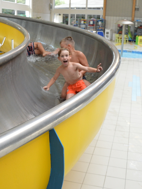 Zwei Kinder rutschen fröhlich auf einer Wasserrutsche in einem Schwimmbad, umgeben von hellen Fliesen und Pflanzen.