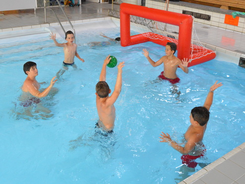 Fünf Jungen spielen im Schwimmbecken Wasserball vor einem roten Tor, alle sind schwimmend oder stehend im Wasser.
