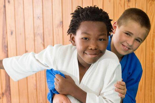 Zwei Jungen in Judogewändern umarmen sich lächelnd auf einer Judomatte vor einer Holzwand.