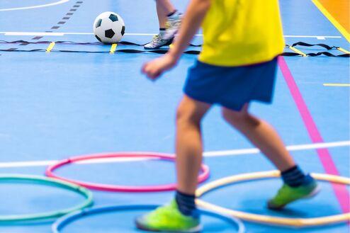 Kinder spielen mit einem Fußball in einer Sporthalle, umgeben von Hula-Hoops auf dem blauen Boden.