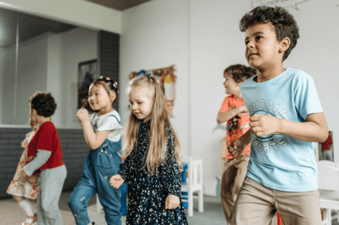 Sechs Kinder tanzen und lachen in einem hellen Raum mit bunten Dekorationen und Spielmöbeln.