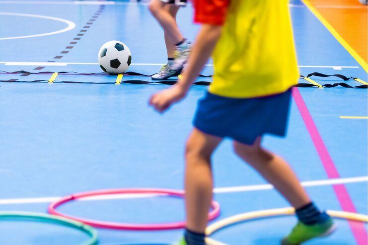 Kinder spielen mit einem Fußball in einer Sporthalle, umgeben von Hula-Hoops auf dem blauen Boden.