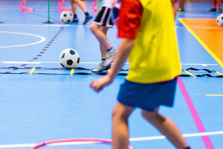 Kinder spielen mit einem Fußball in einer Sporthalle, umgeben von Hula-Hoops auf dem blauen Boden.