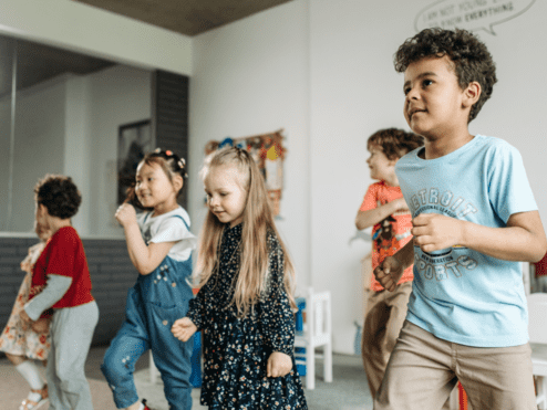 Sechs Kinder tanzen und lachen in einem hellen Raum mit bunten Dekorationen und Spielmöbeln.