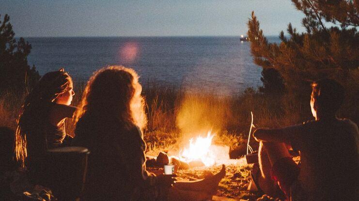 Freundeskreis sitzt am Lagerfeuer am Strand, Blick auf das Meer bei Nacht, umgeben von Bäumen und Gras.
