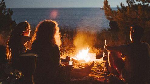 Freundeskreis sitzt am Lagerfeuer am Strand, Blick auf das Meer bei Nacht, umgeben von Bäumen und Gras.