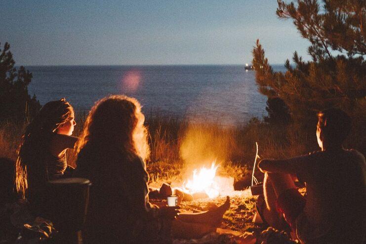 Freundeskreis sitzt am Lagerfeuer am Strand, Blick auf das Meer bei Nacht, umgeben von Bäumen und Gras.