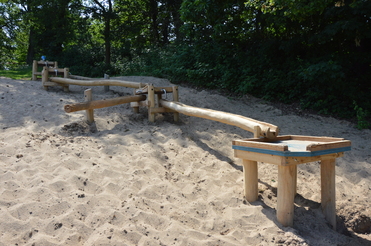 Holzspielgerät auf Sandboden, bestehend aus langen, gebogenen Balken und einem kleinen Tisch mit Sandfläche.