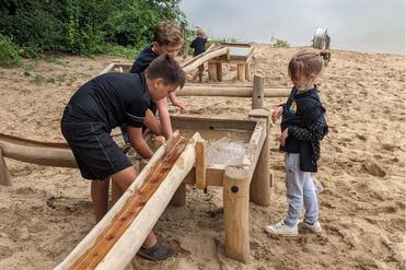 Kinder spielen an einem Wasserspielplatz mit Holzstrukturen und Sand, während im Hintergrund ein Gewässer zu sehen ist.