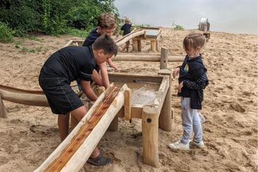 Kinder spielen an einem Wasserspielplatz mit Holzstrukturen und Sand, während im Hintergrund ein Gewässer zu sehen ist.