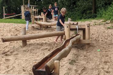 Kinder spielen an einem Wasserspielplatz mit Holzrutschen und sandigem Untergrund.