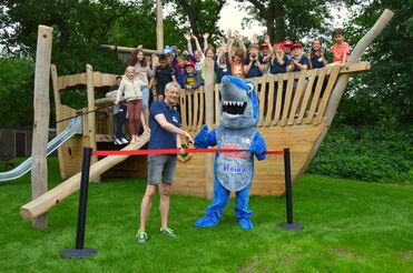 Eröffnung eines Spielplatzes mit einem Mann und einem Hai-Maskottchen, während Kinder auf einem Piratenschiff jubeln.