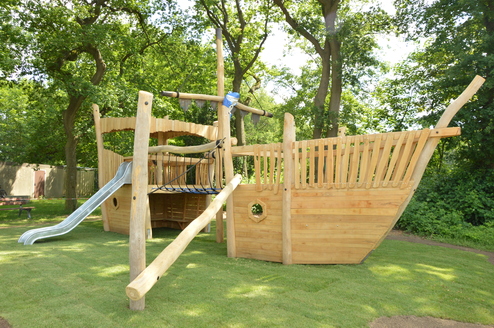 Holzspielschiff mit Rutsche und Kletterseil in einem Spielgarten, umgeben von Bäumen und grünem Rasen.
