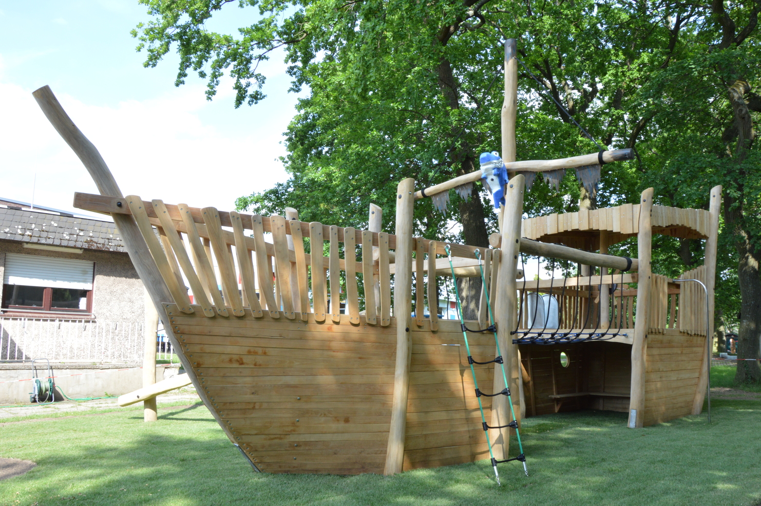 Holzspielplatz in Form eines Schiffes mit Rampen, Spielm&ouml;glichkeiten und einer gr&uuml;nen Rasenfl&auml;che im Hintergrund.