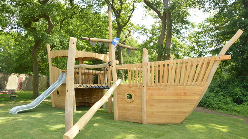 Holzspielschiff mit Rutsche und Kletterseil in einem Spielgarten, umgeben von Bäumen und grünem Rasen.