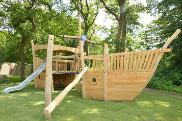 Holzspielschiff mit Rutsche und Kletterseil in einem Spielgarten, umgeben von Bäumen und grünem Rasen.