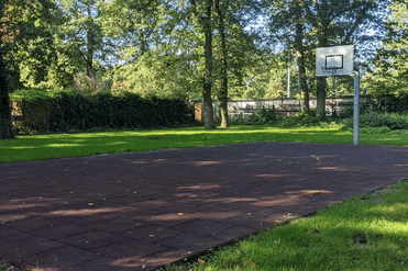 Basketballplatz mit Gummiboden und Korb, umgeben von Rasen und Bäumen in einem Park.