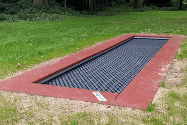 Rechteckiger Trampolinrahmen mit schwarzer Sprungfläche, umgeben von grünem Gras und Bäumen im Hintergrund.