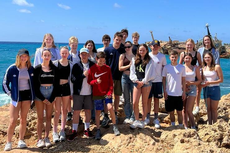 Gruppe von Jugendlichen steht auf Felsen am Meer, im Hintergrund ein Schiff und blauer Himmel.