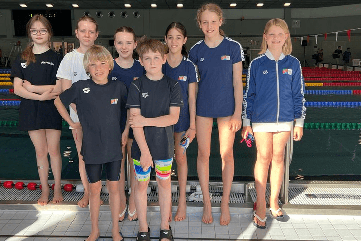 Gruppe von acht Schwimmern in Vereinskleidung steht am Schwimmbeckenrand, lächelnd in einer Sporthalle.