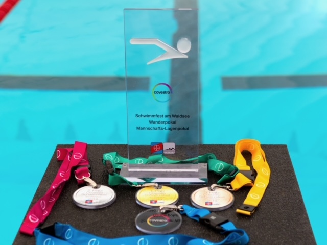 Schwimmmedaillen und ein Pokal liegen auf einem Tisch vor einem Schwimmbecken mit blauen Wasserbahnen.