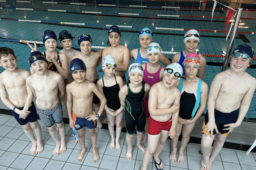 Gruppe von 16 schwimmenden Kindern in Badekleidung und Schwimmhauben am Poolrand.