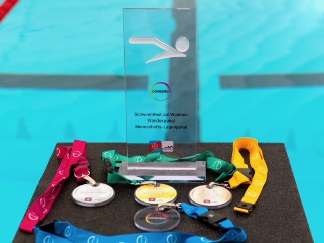 Schwimmmedaillen und ein Pokal liegen auf einem Tisch vor einem Schwimmbecken mit blauen Wasserbahnen.