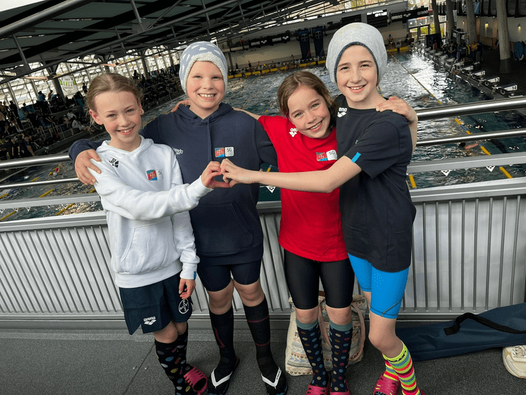 Vier Kinder in Schwimmbekleidung machen ein Herz mit den Händen auf einem Schwimmbecken-Hintergrund.