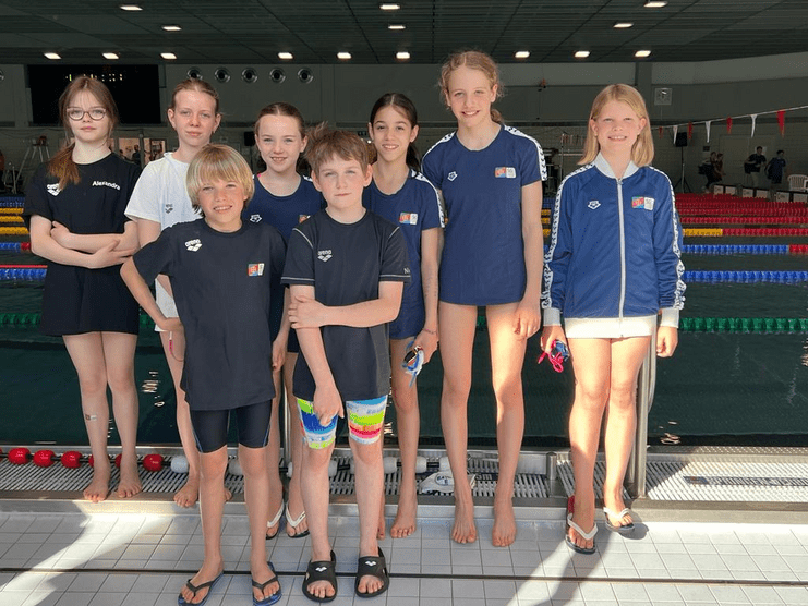 Gruppe von acht Schwimmern in Vereinskleidung steht am Schwimmbeckenrand, lächelnd in einer Sporthalle.