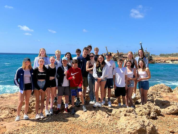 Gruppe von Jugendlichen steht auf Felsen am Meer, im Hintergrund ein Schiff und blauer Himmel.