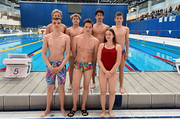 Gruppe von Schwimmern in Badebekleidung steht am Beckenrand eines Schwimmbeckens mit Wettkampfanlagen.