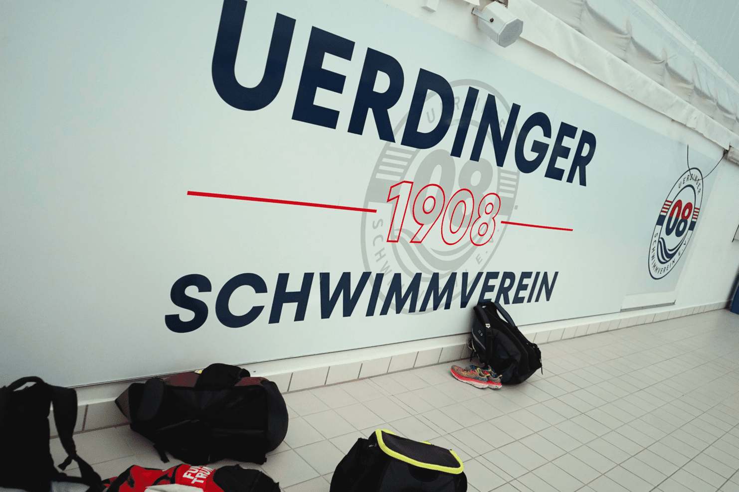 Wand mit dem Schriftzug "Uerdinger Schwimmverein 1908" und mehreren Sporttaschen auf dem Boden.