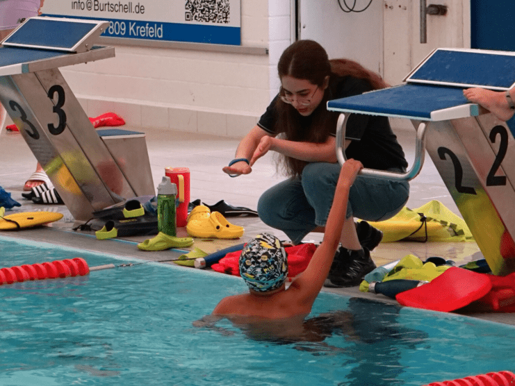 Eine Betreuerin hilft einem schwimmenden Kind beim Verlassen des Beckens an der Startblöcken im Schwimmbad.