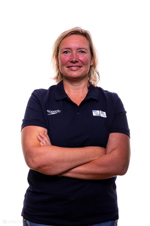 Porträt einer lächelnden Frau mit blondem Haar in einem dunkelblauen Poloshirt, die vor weißem Hintergrund steht.