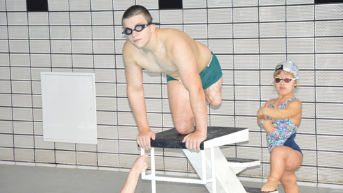 Junger Schwimmer in Schwimmbrille springt vom Startblock ins Wasser, während ein Mädchen im Badeanzug zuschaut.