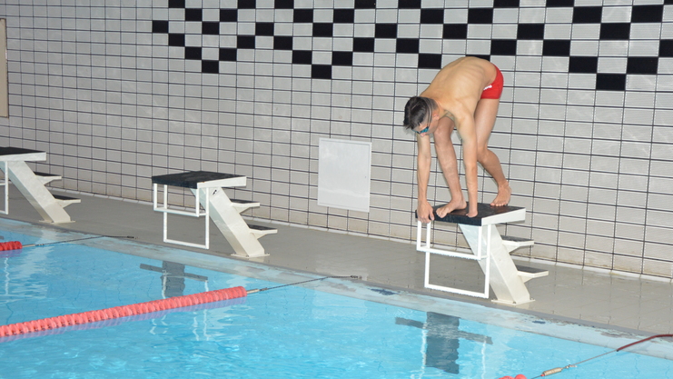 Ein Schwimmer in roten Badehosen steht bereit auf einem Startblock neben einem blauen Schwimmbecken.