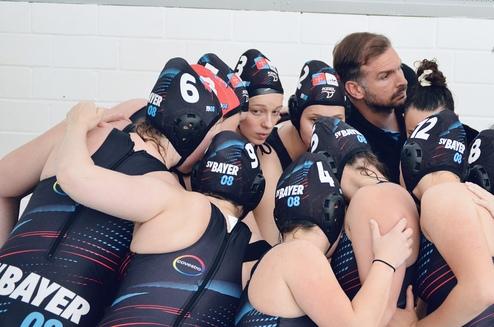 Wasserball-Team versammelt sich im Huddle, Trainer gibt Anweisungen, Spielerinnen tragen schwarze und rote Badekappen.