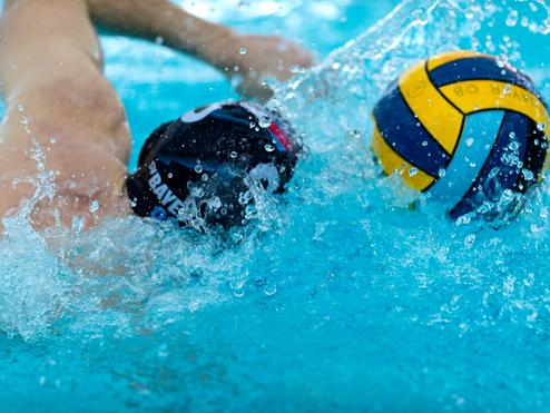 Spritzernde Wassertropfen während eines Wasserball-Spiels mit zwei Spielern und einem Ball im Fokus.