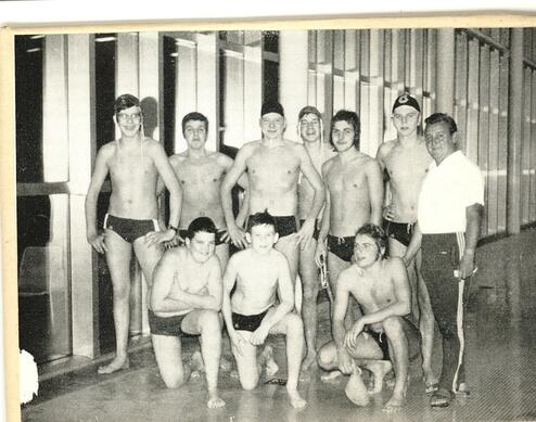 Gruppe von Jungen und einem Trainer in Badehosen, lächelnd vor einer Schwimmbadwand mit großen Fenstern.