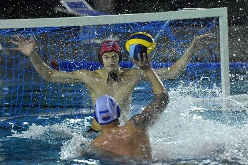 Wasserballspieler blockiert einen Wurf mit einem Ball in einem spritzenden Wasserbecken.