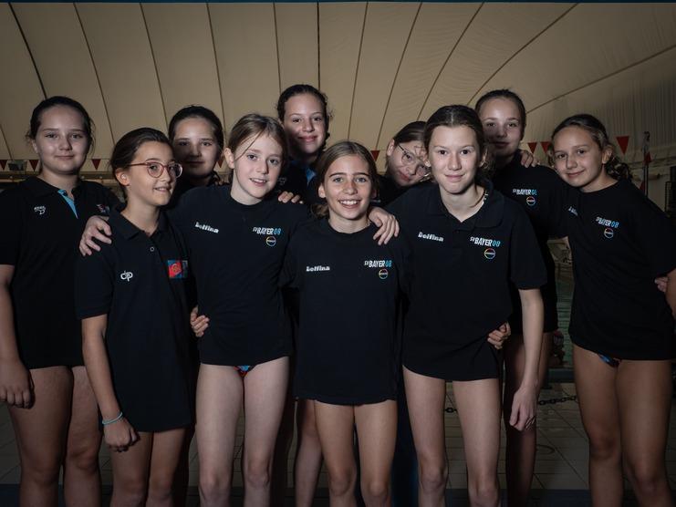 Gruppenfoto von zehn jungen Schwimmerinnen in schwarzen T-Shirts vor einem Schwimmbecken.