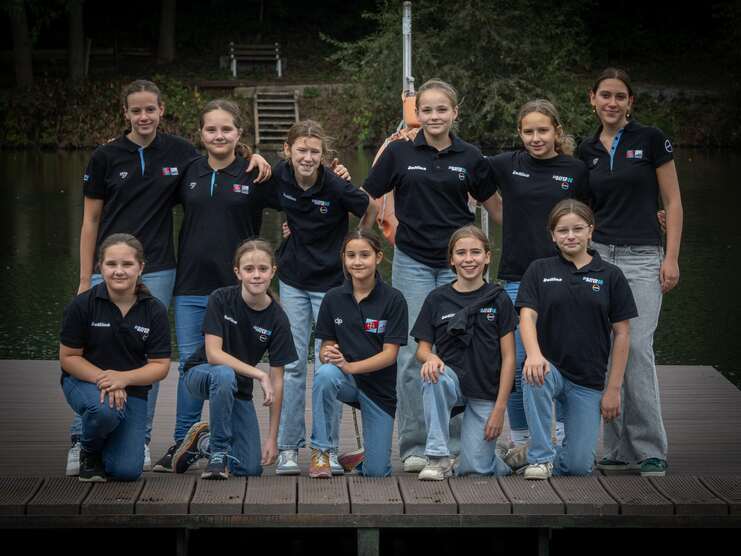 Gruppe von zwölf Mädchen in schwarzen Poloshirts, stehend und sitzend auf einem Holzsteg am Wasser.