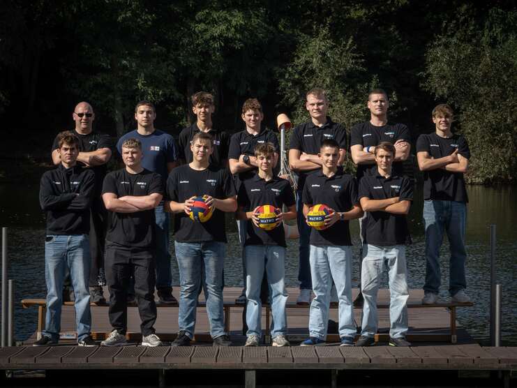 Gruppenbild von fünfzehn Sportlern in schwarzen T-Shirts, die Wasserbälle halten, auf einem Steg am Ufer eines Gewässers.
