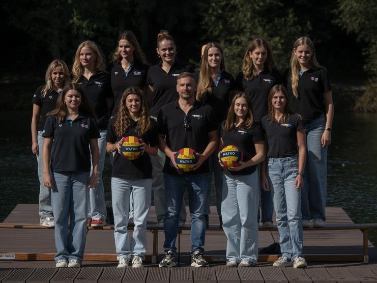 Gruppe von zwölf Frauen und einem Mann in schwarzen Shirts, die Wasserbälle auf einem Steg vor einem Gewässer halten.