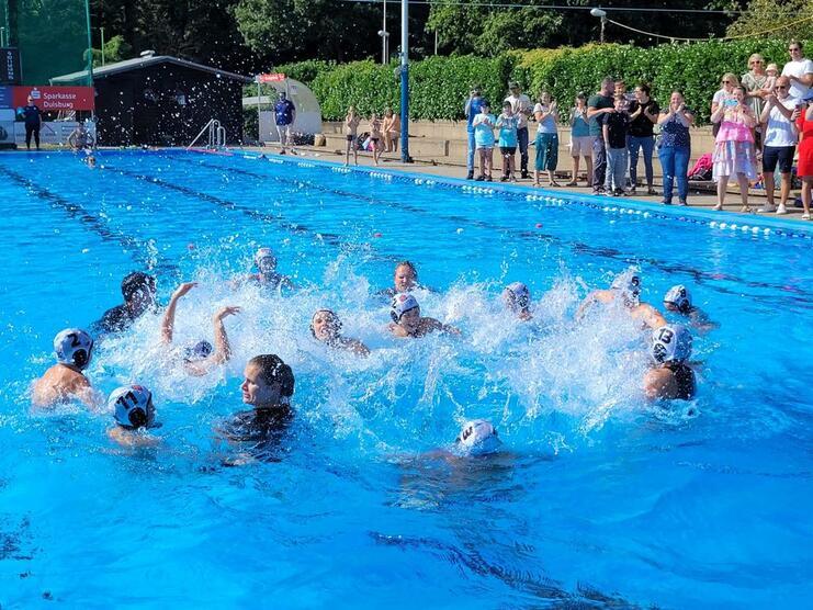 Wasserspiele von mehreren Spielern in einem Schwimmbecken, umgeben von zuschauenden Personen am Beckenrand.